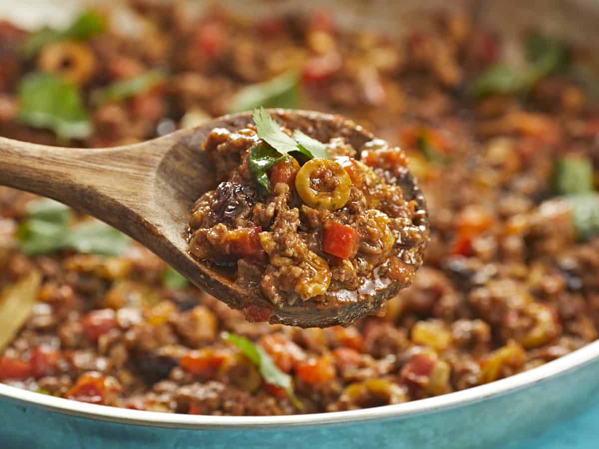 Wooden spoon full og picadillo in the foreground with picadillo in a silver skillet in the background.