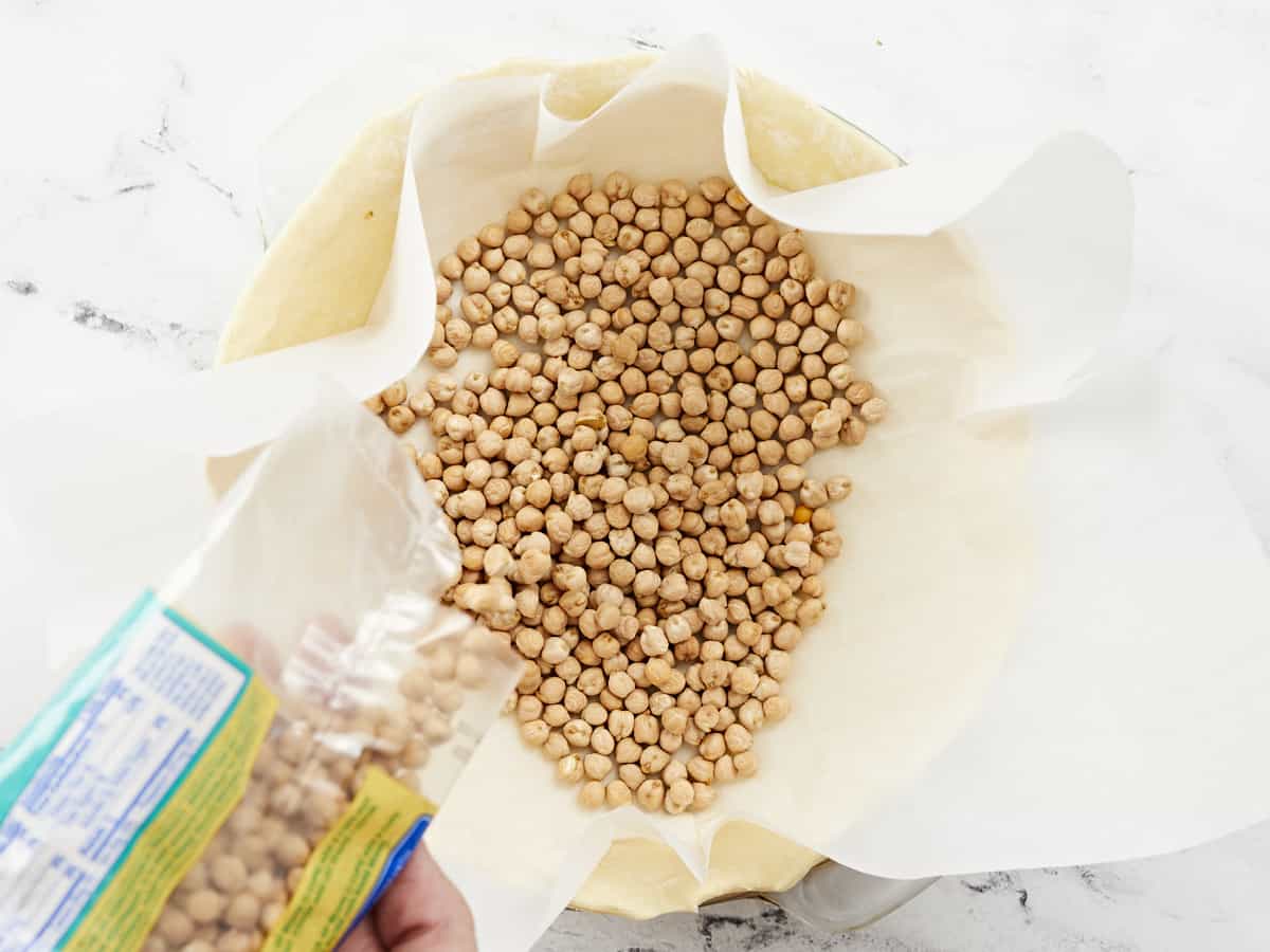 Overhead shot of dry beans/ pie weights being added to the parchment paper-lined sheet of puff pastry.