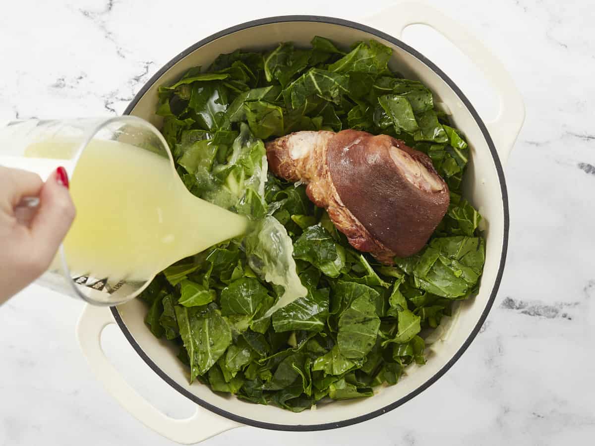 Collard greens and ham hock in the pot, chicken broth being poured in.