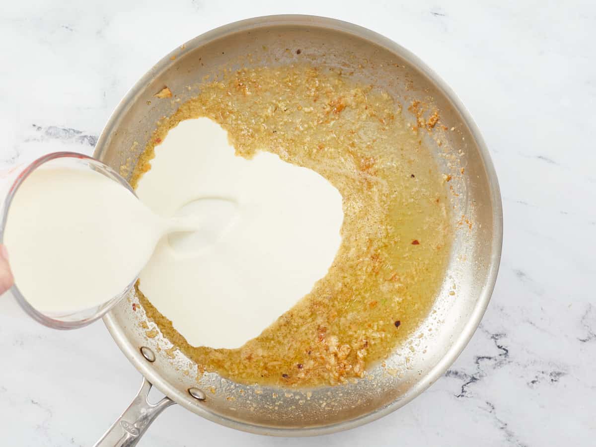 A large stainless steel skillet with butter and sautéed garlic, and heavy cream being poured from a glass measuring cup into the skillet from the left side of the pan.