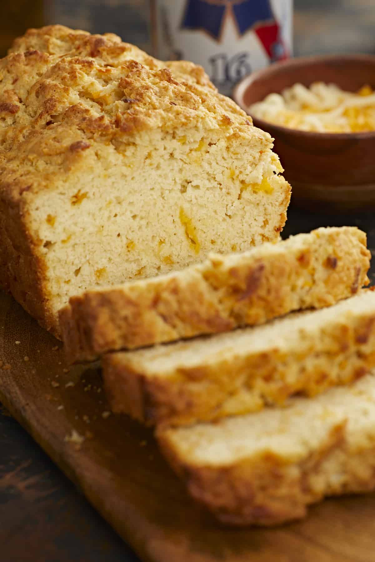 A vertical close up of a loaf of bread on a wooden cutting board with three slices slightly overlapping in the foreground of the image, and behind the cutting board is a small wooden bowl filled with shredded cheese and a can of beer is visible in the upper right corner of the frame.