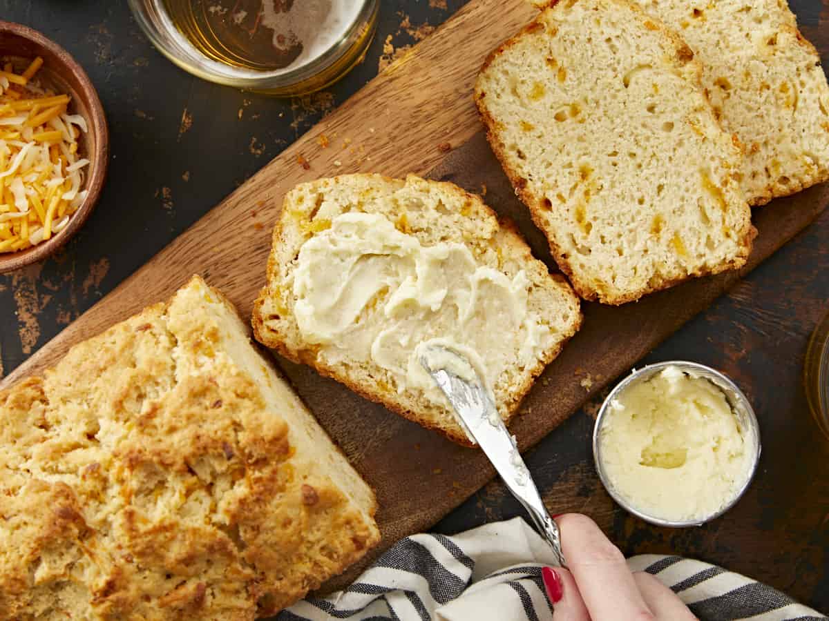 A close up shot of long wooden cutting board with a half a loaf of beer bread in the left corner of the frame, two slices on the upper right corner of the frame and in between a slice is being covered with salted homemade butter with a small metal butter knife. 