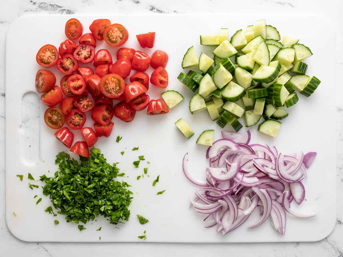 Prepped tomatoes, cucumbers, onions, and parsley on a cutting board.