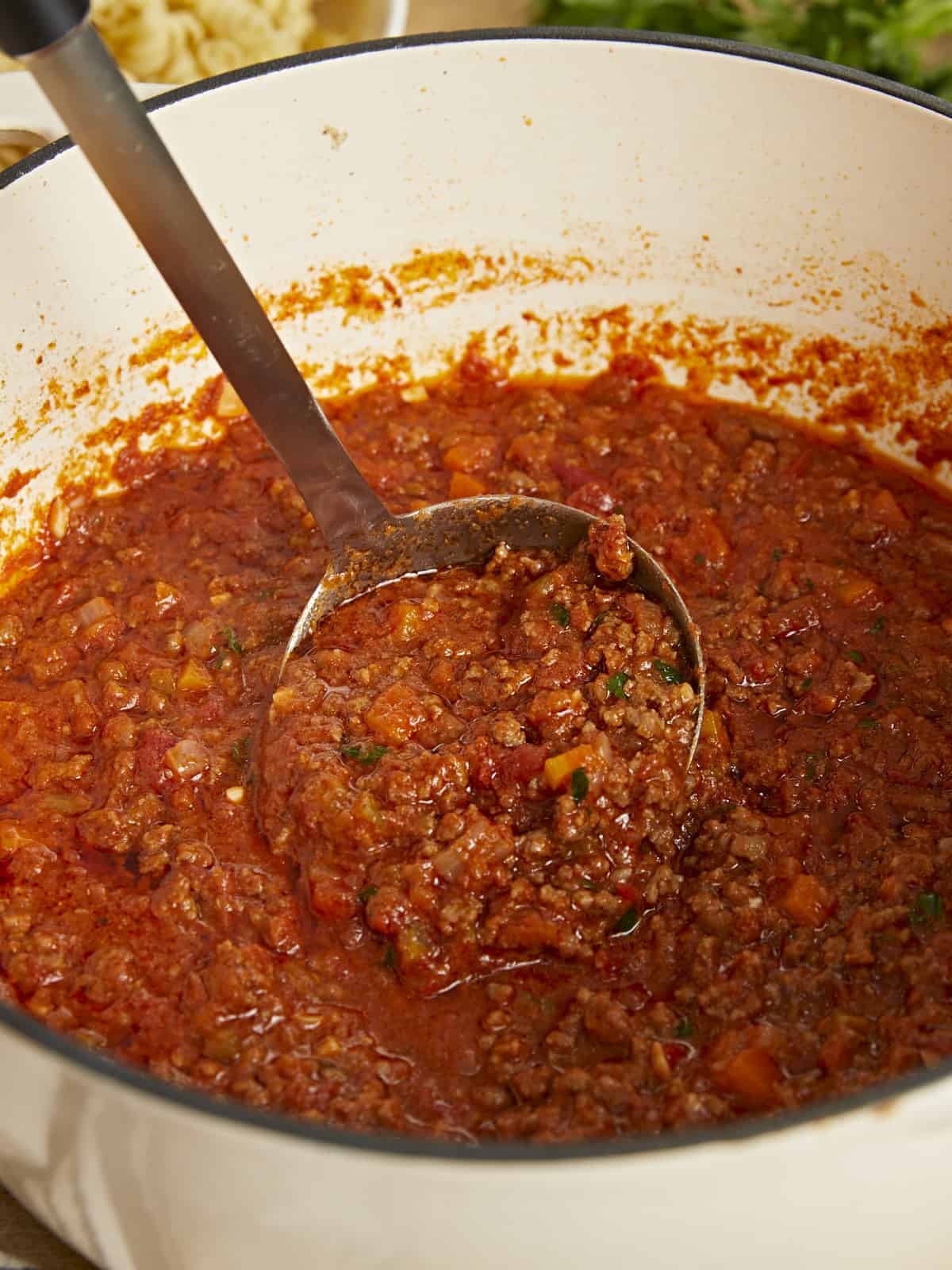 Side shot of bolognese in a beige Dutch oven with a metal spoon in it.