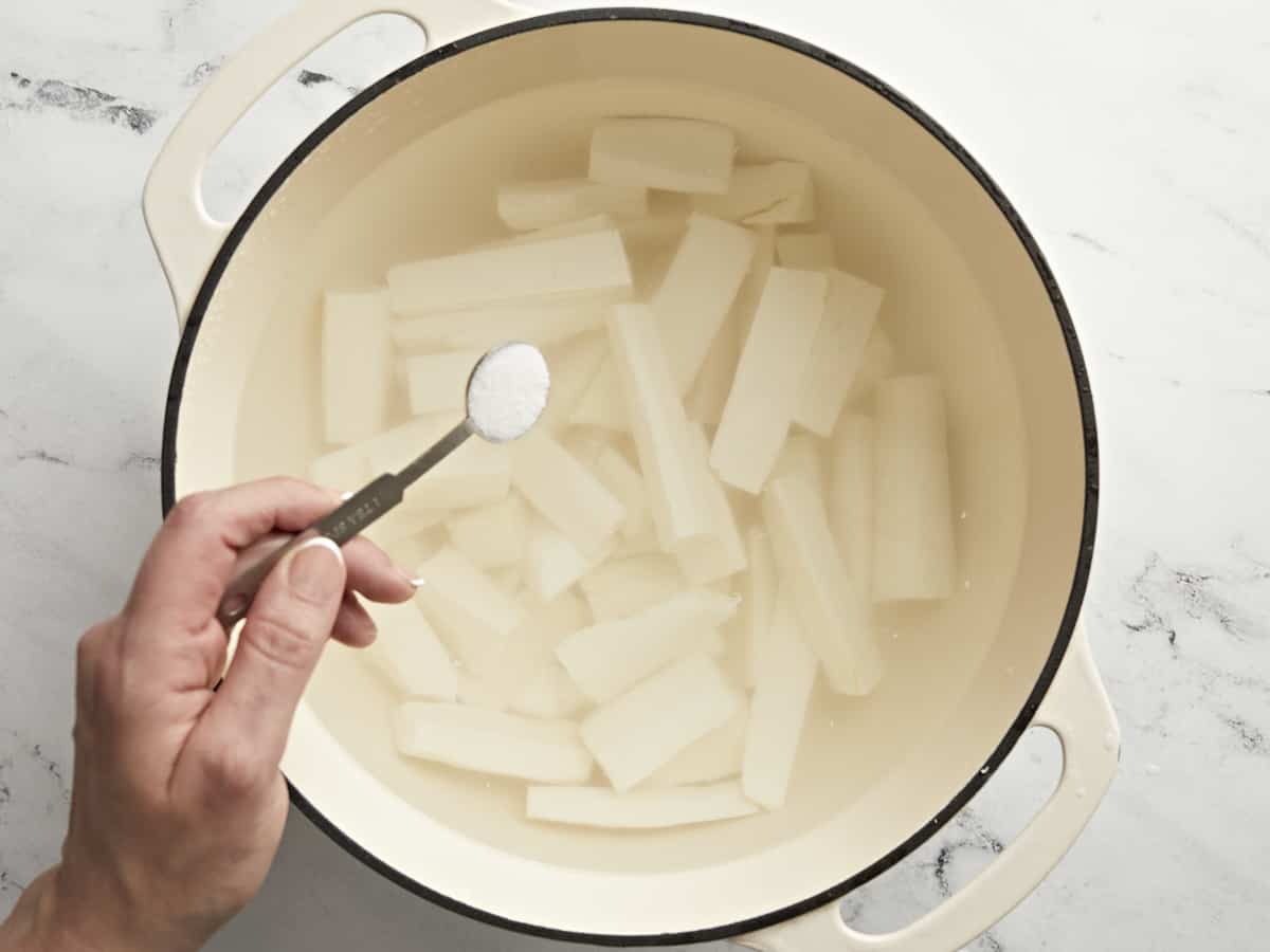 Overhead shot of yuca in a large cream-colored pot with salt being added to it.