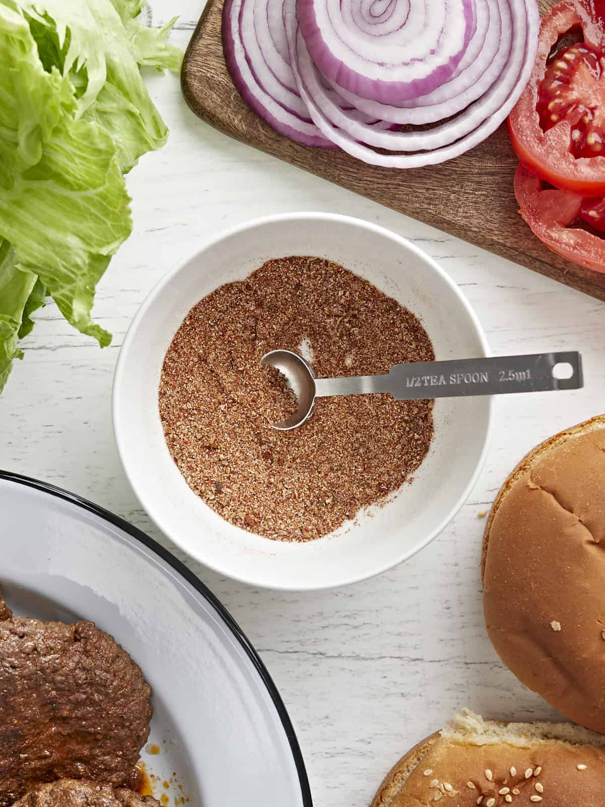 Overhead view of a bowl full of burger seasoning surrounded by burger toppings.