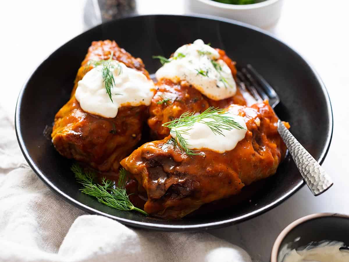 Side view of cabbage rolls served on a black plate with sour cream and fresh dill on top and a fork on the side.