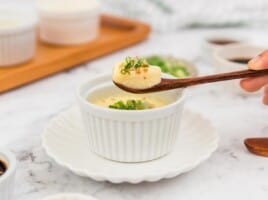 Steamed eggs in a white ramekin garnished with green onions and a spoon lifting some eggs out of the ramekin.