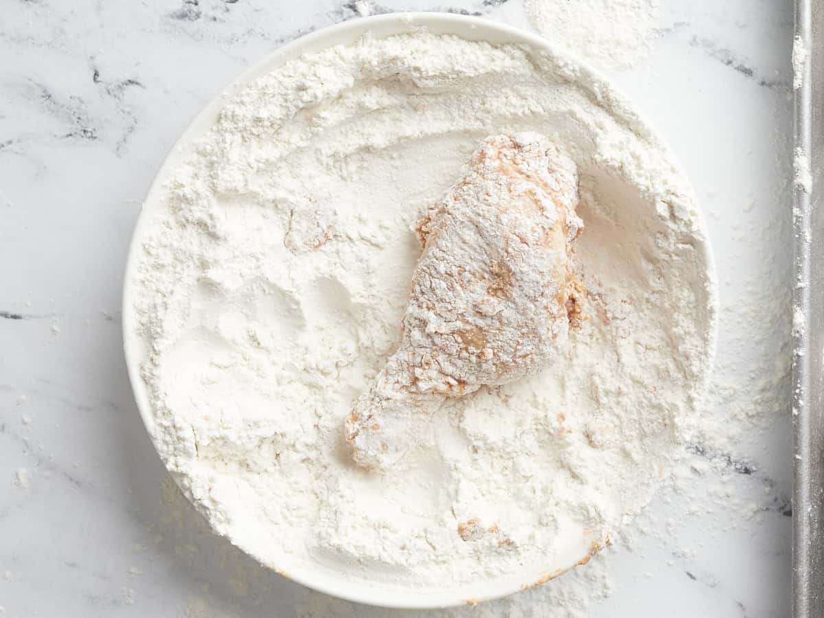 Overhead shot of brined chicken being placed in a white bowl full of flour.