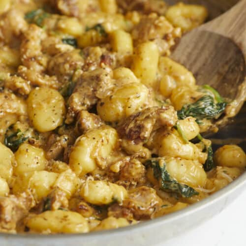 Side shot of Creamy Chicken and Gnocchi in a silver skillet with a wooden spoon in it.