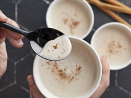 Overhead shot of three white bowls of Maizena with cinnamon sprinkled on top with one bowl one the forground being held by a hand and with a black spoon being dipped into it.