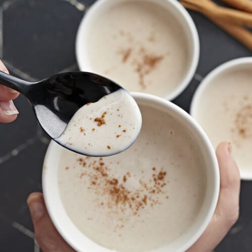 Overhead shot of three white bowls of Maizena with cinnamon sprinkled on top with one bowl one the forground being held by a hand and with a black spoon being dipped into it.