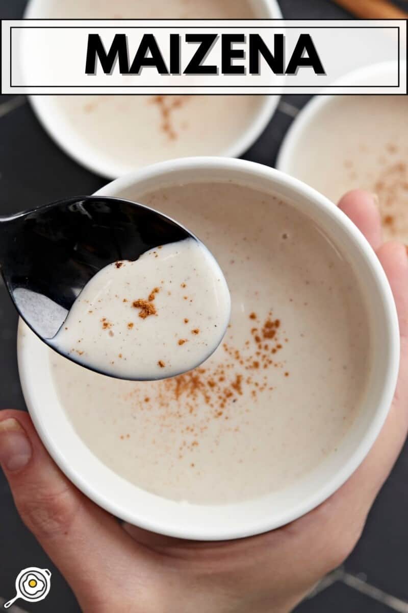 Overhead shot of three white bowls of Maizena with cinnamon sprinkled on top with one bowl in the foreground being held by a hand and with a black spoon being dipped into it.