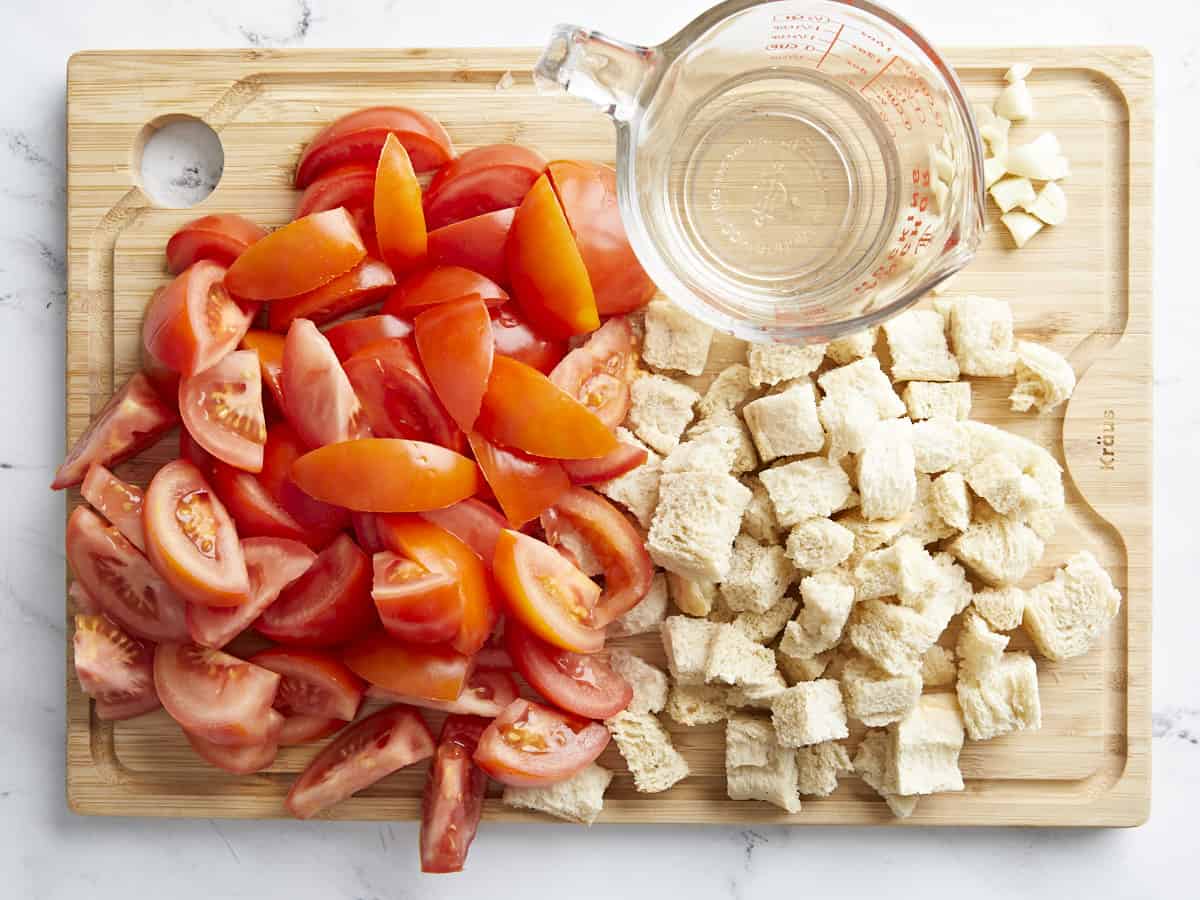 Overhead shot of Salmorejo's main ingredients: chopped fresh tomato, chopped day-old bread, and water, on a wood cutting board.