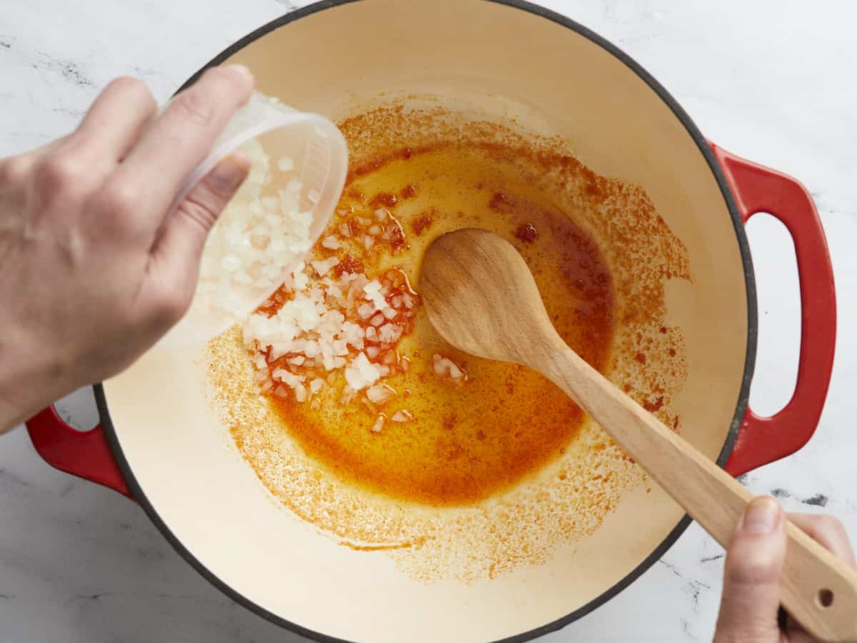 Overhead shot of a hand adding onion to a red Dutch oven while a second hand stirs with a wooden spoon.