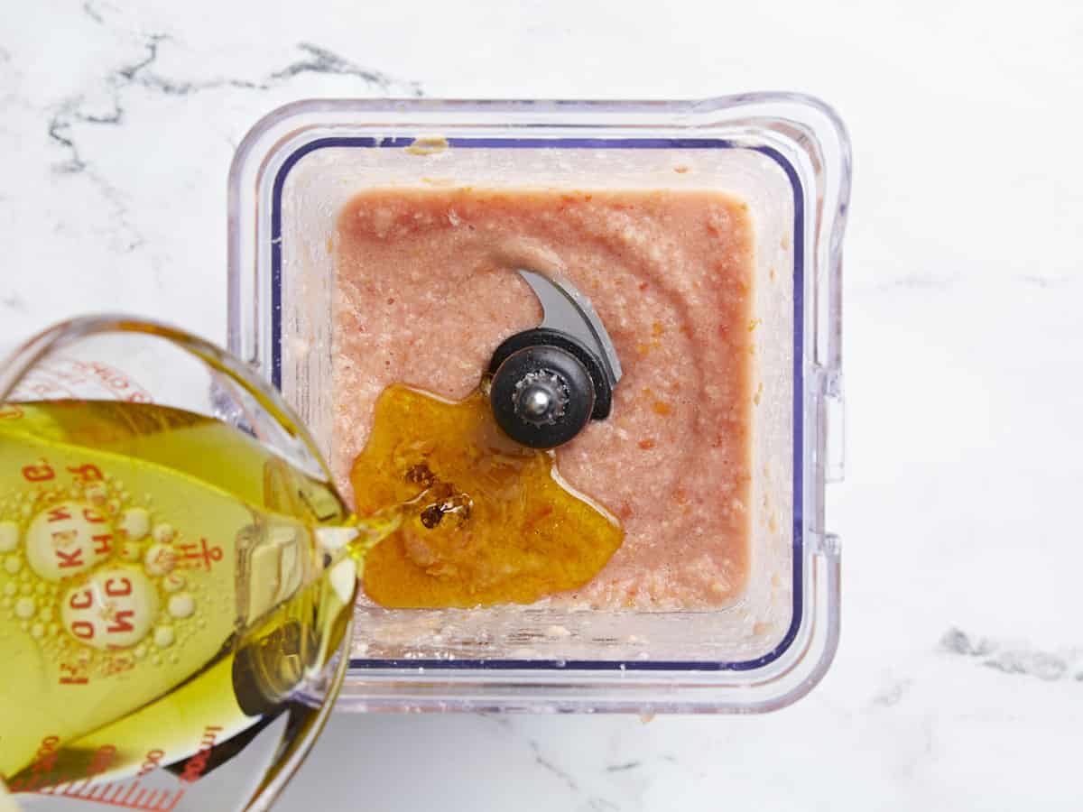 Overhead shot of olive oil being drizzled into a blender jar full of pureed tomato and bread.