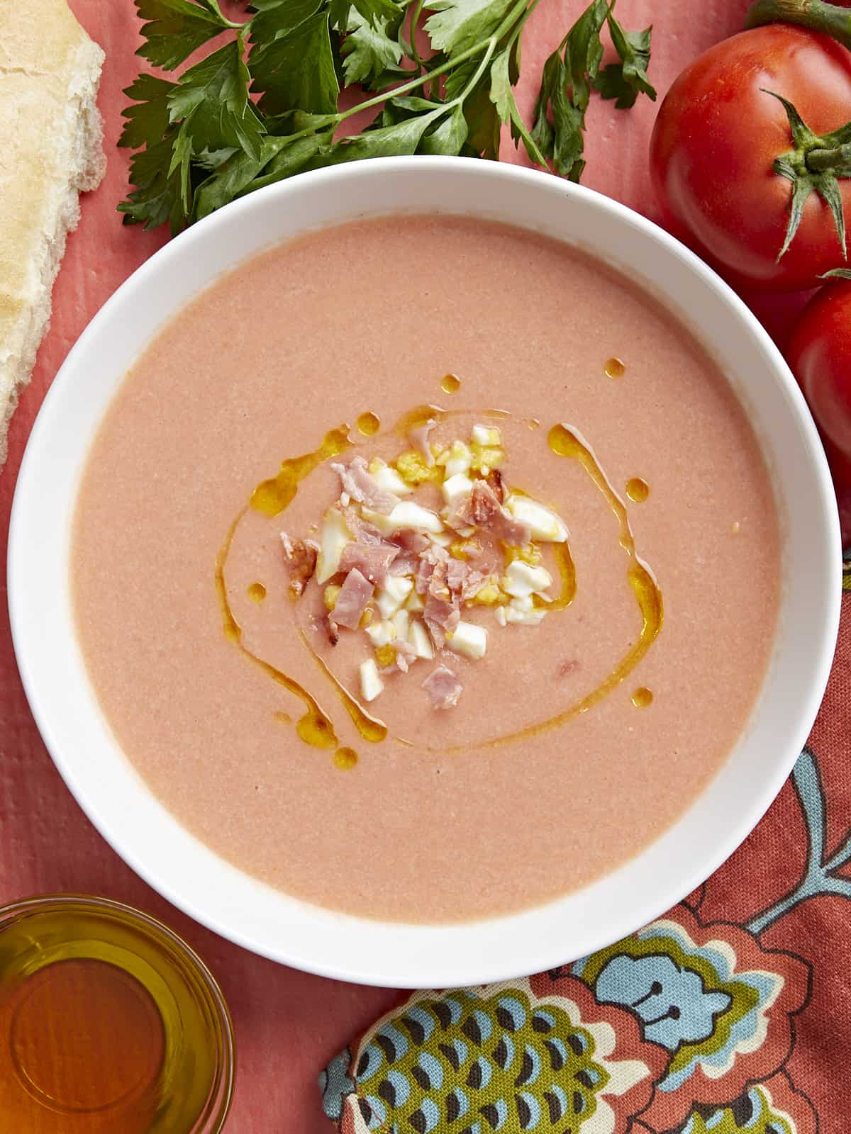 Overhead shot of a white bowl of Salmorejo garnished with a drizzle of olive oil, chopped hard-boiled eggs, and crispy ham crumbles. 