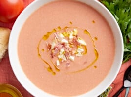 Overhead shot of a white bowl of Salmorejo garnished with a drizzle of olive oil, chopped hard-boiled eggs, and crispy ham crumbles.