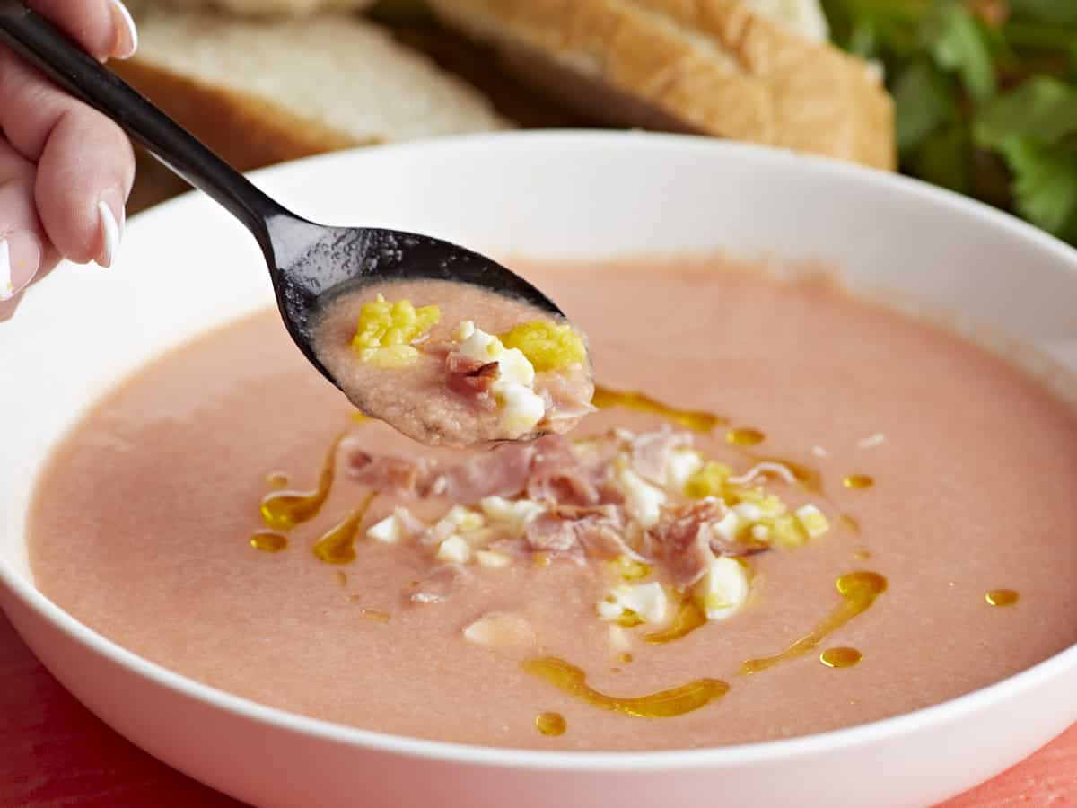 Side shot of a white bowl of Salmorejo garnished with a drizzle of olive oil, chopped hard-boiled eggs, and crispy ham crumbles with a black spoon coming out of it.