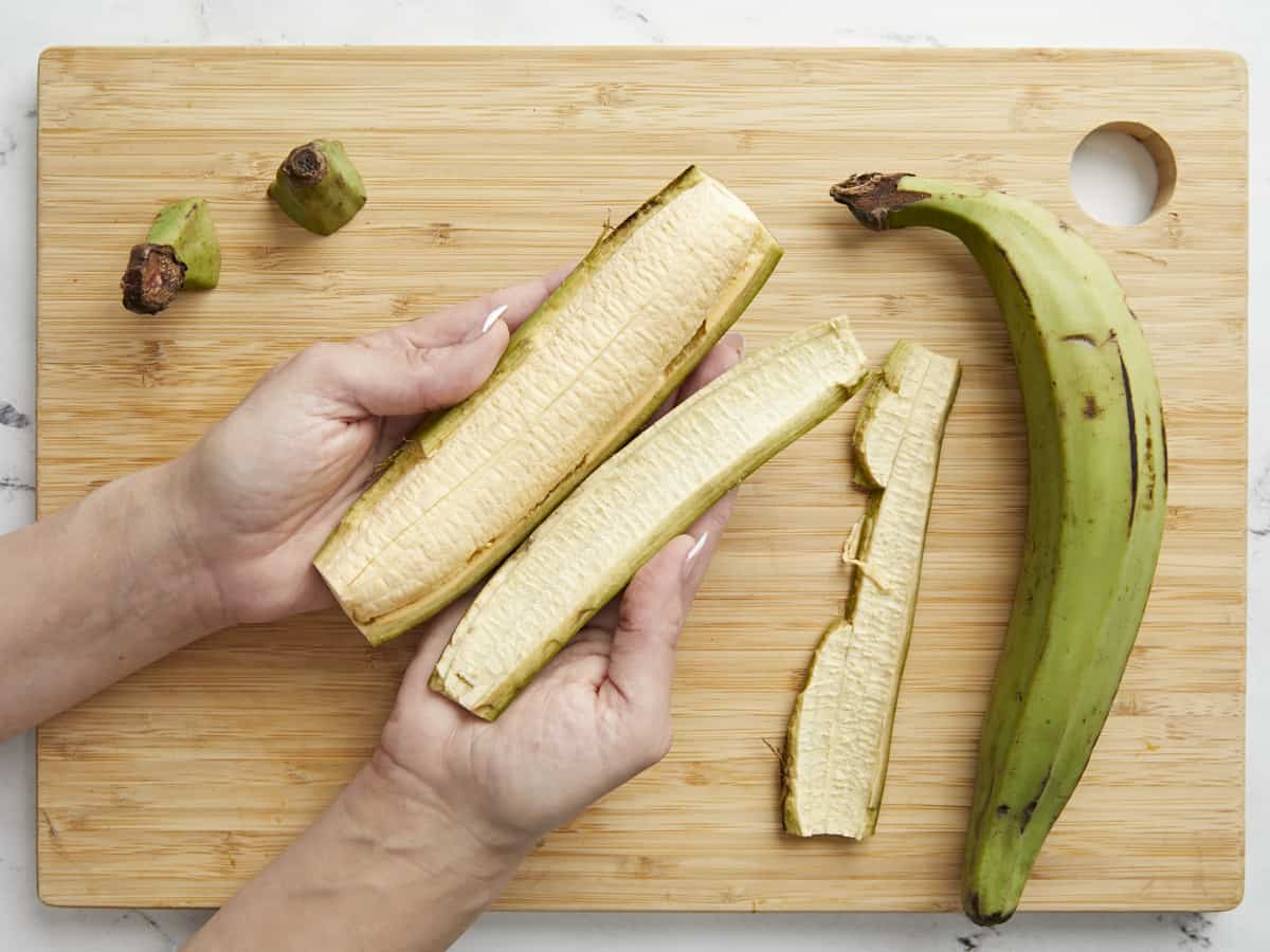 Overhead shot of green plantain being peeled on a wood cutting board.