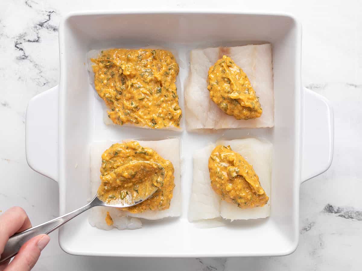 Garlic butter being spread over fish filets in a baking dish.