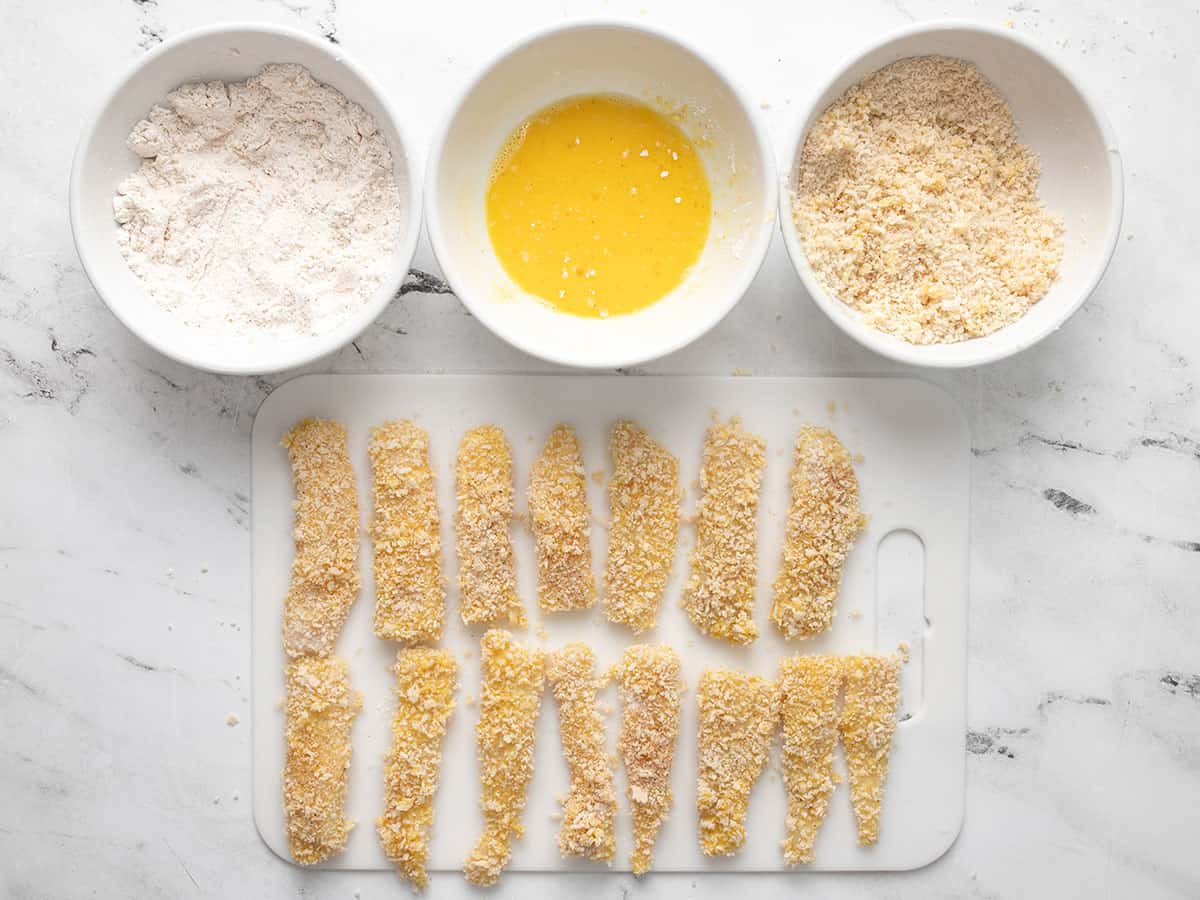 Breaded fish sticks on a cutting board next to the three bowls of ingredients.