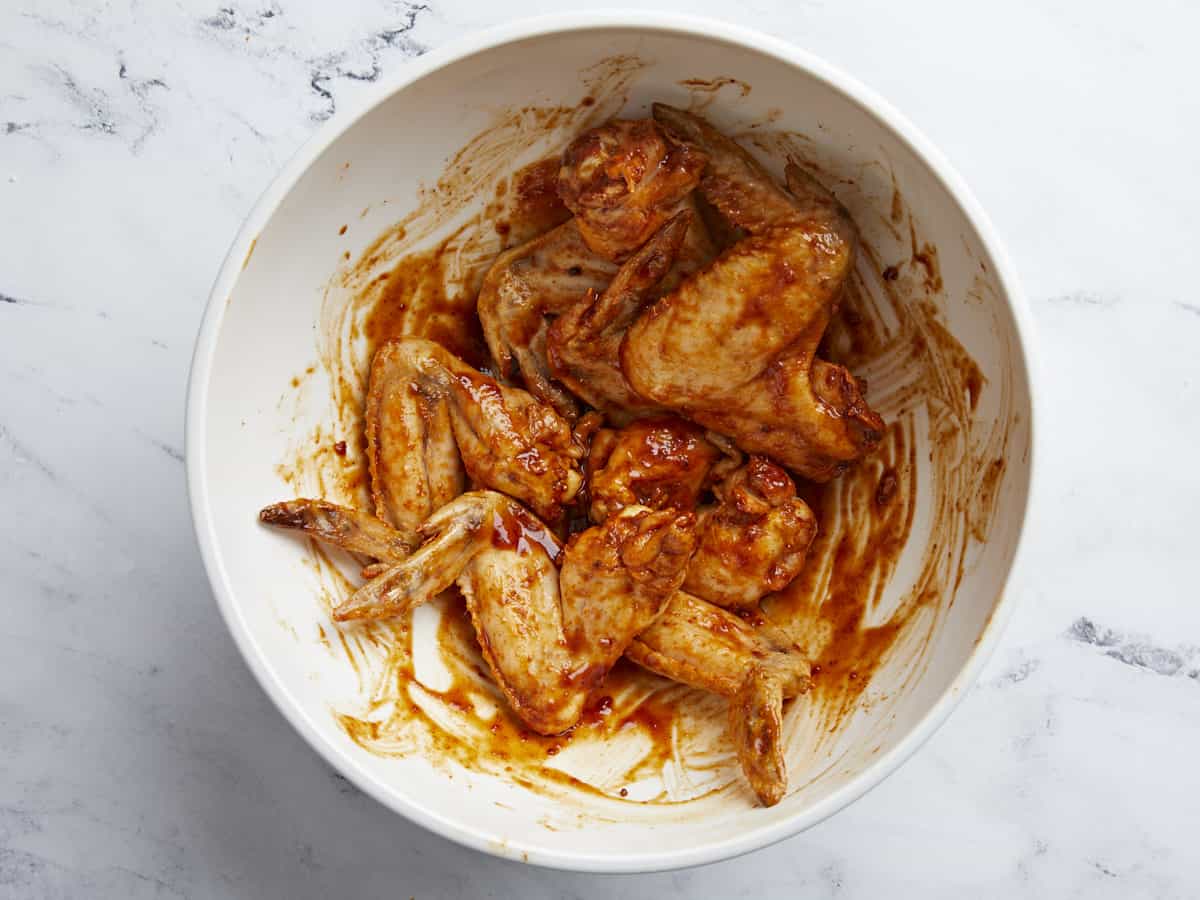 Overhead shot of chicken wings being mixed into BBQ sauce in a white bowl.