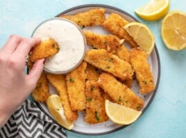 A hand dipping a fish stick into a bowl of tartar sauce.