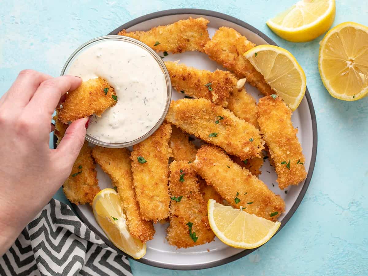 A hand dipping a fish stick into a bowl of tartar sauce.