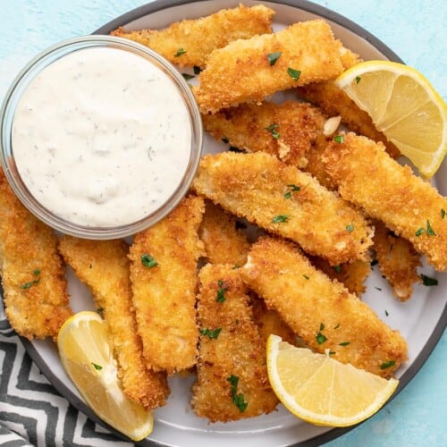 Overhead view of a plate full of fish sticks with tartar sauce and lemon wedges.