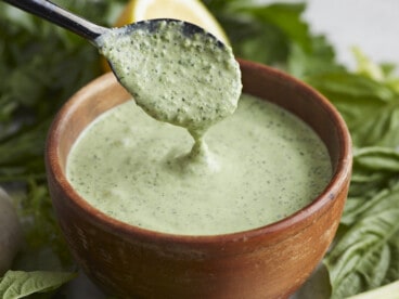 Side shot of Green Goddess Dressing in a wood bowl with a black spoon drizzling some in.