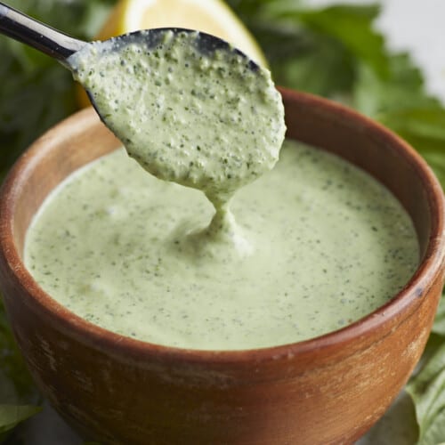 Side shot of Green Goddess Dressing in a wood bowl with a black spoon drizzling some in.