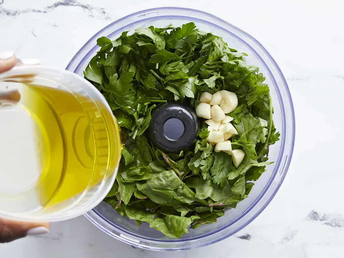 Swiss chard, Spinach, parsley, cilantro, and garlic added to a food processor with olive oil being poured in.