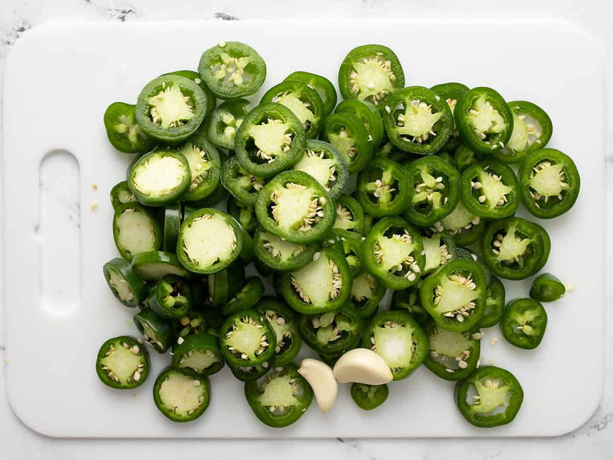 Sliced jalapeños and peeled garlic on a cutting board.