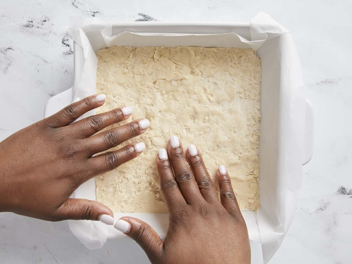 Shortbread crust being pressed into a white baking dish.