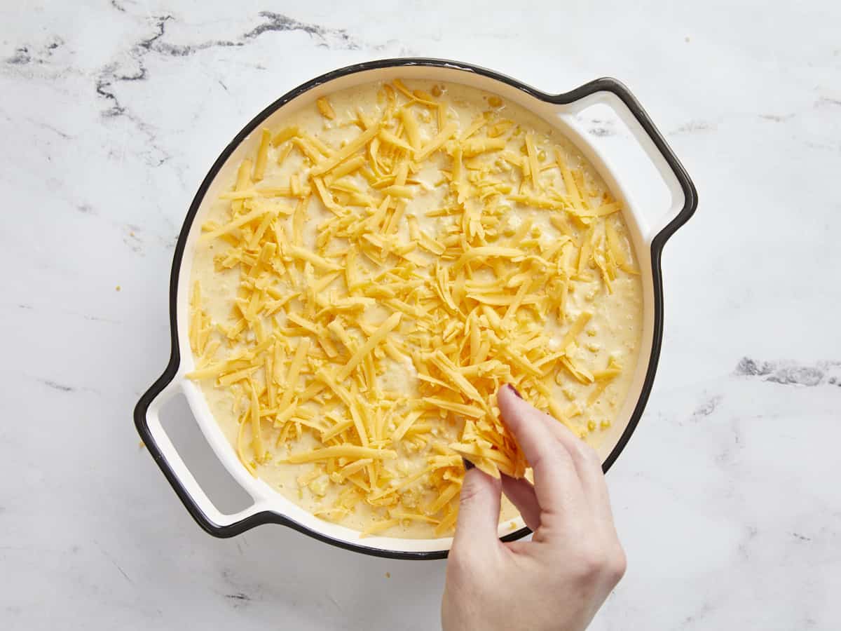 Cheddar cheese being sprinkled over top of the corn pudding in the casserole dish.