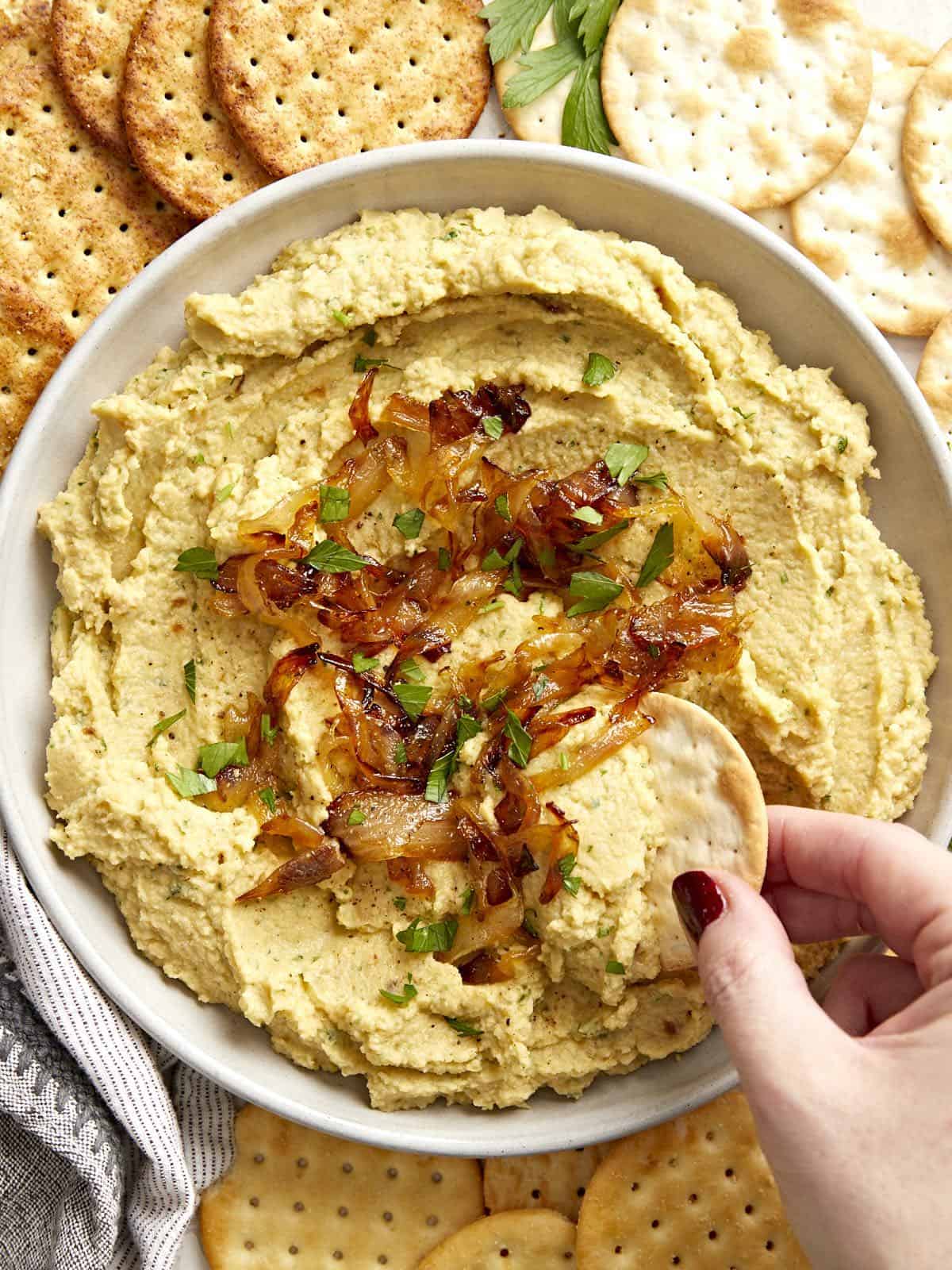 Close up overhead view of a bowl full of chickpea spread with caramelized onions, a cracker being dipped into the side.