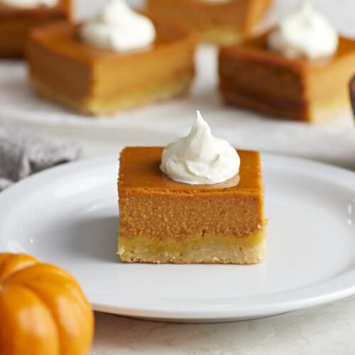 Side view of a pumpkin pie bar on a white plate with whipped cream on top.