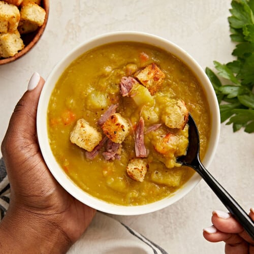 Overhead view of a single bowl of split pea soup with one hand holding the bowl and another hand spooning some of the soup out.