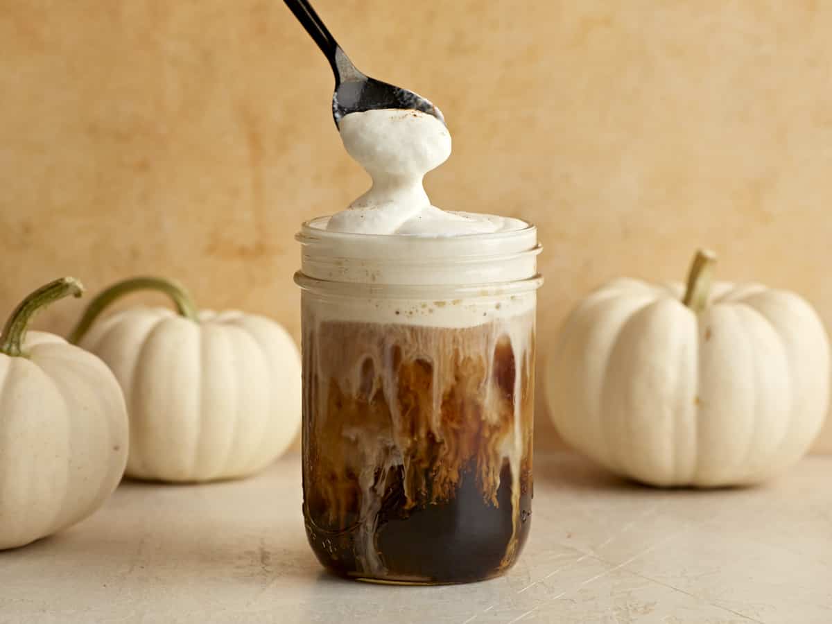 A spoon lifting some sweet cream cold foam off the top of the glass of iced coffee.