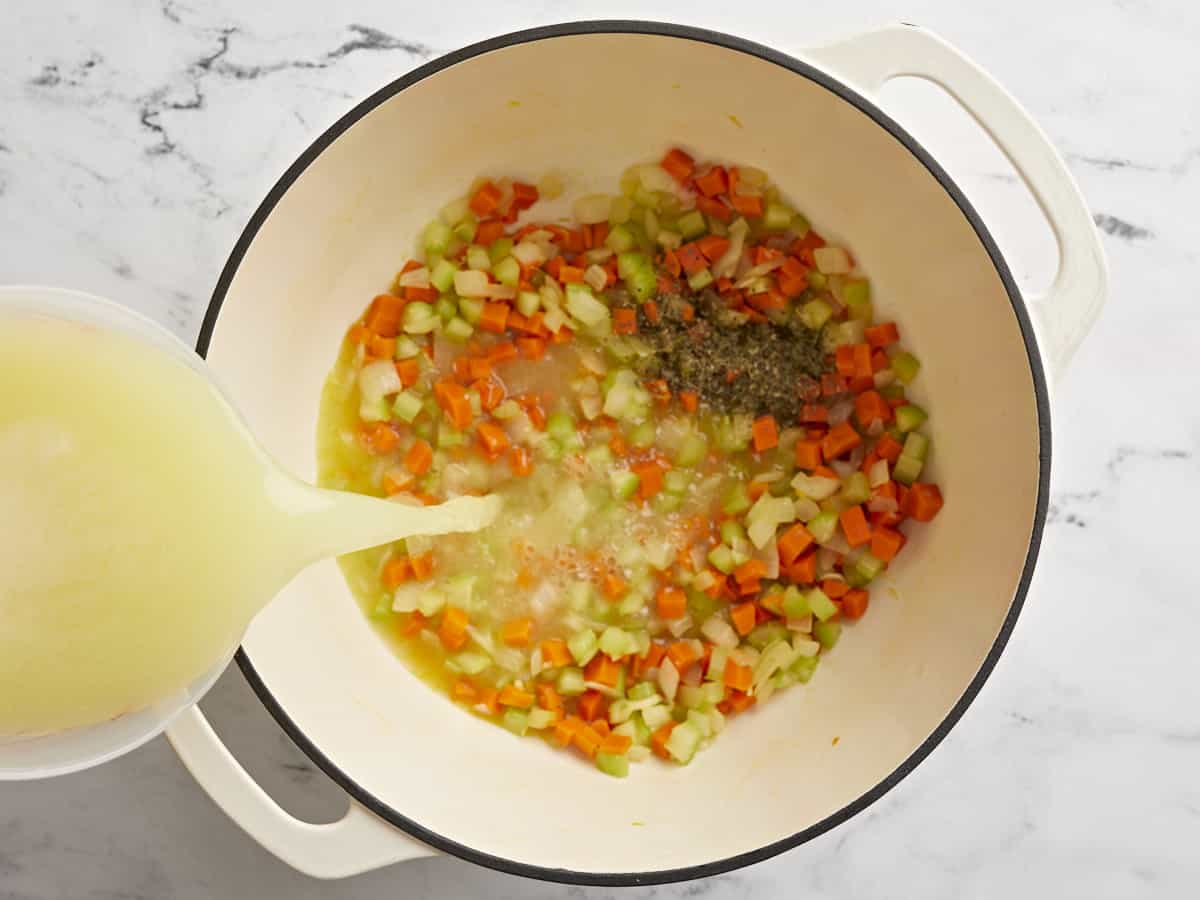 Cooked vegetables in the soup pot with Italian seasoning and chicken broth being poured in.