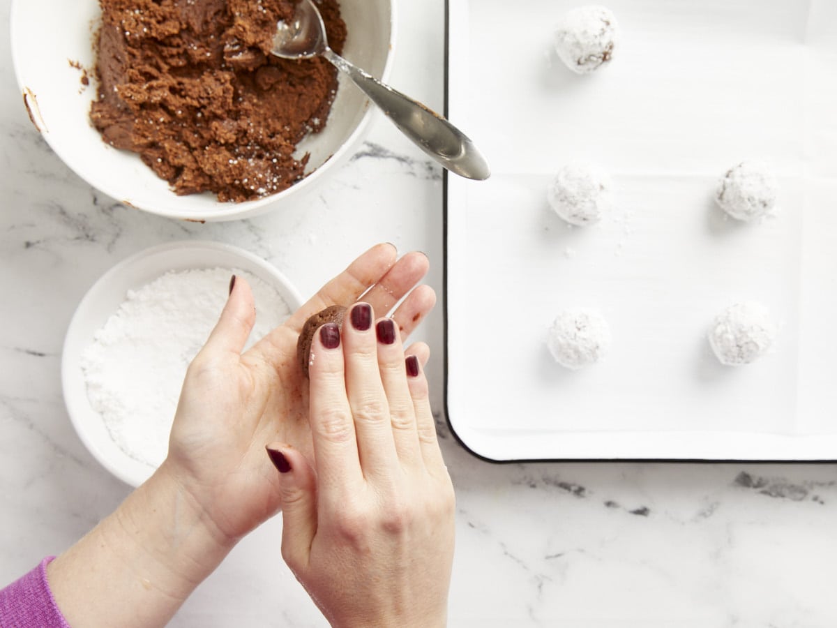 Chocolate crinkle cookies being shaped and rolled in powdered sugar. 