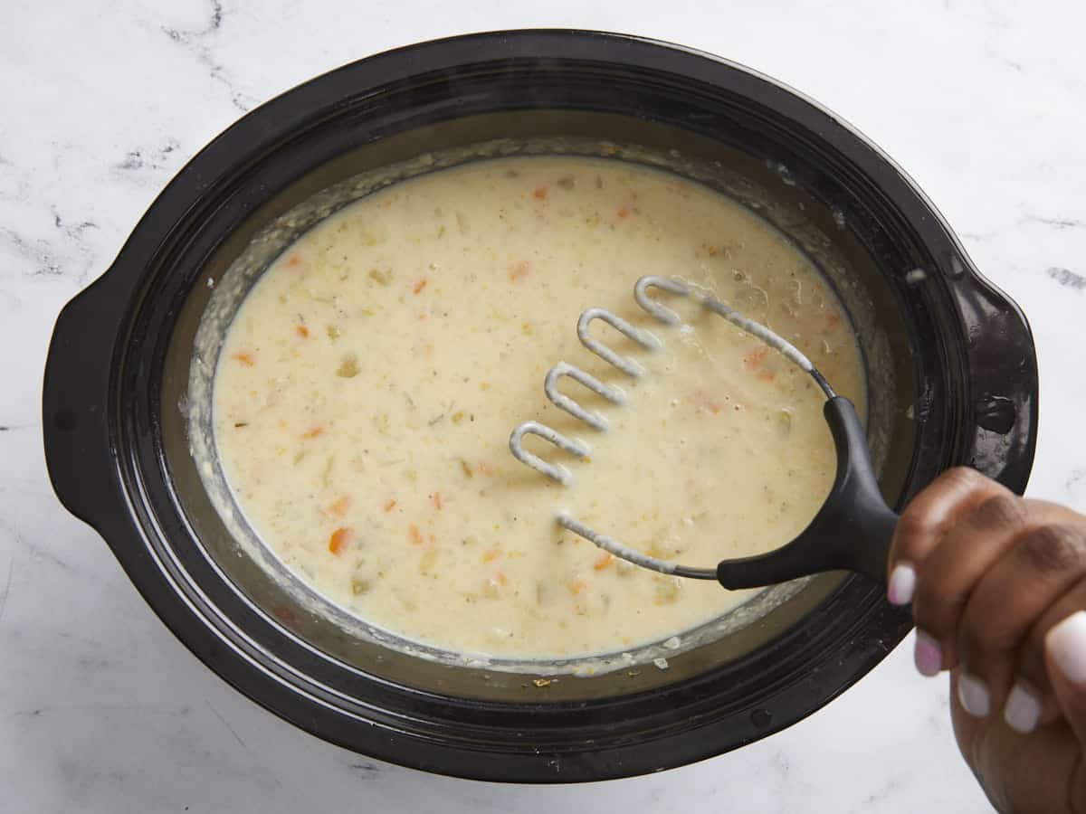 Potato soup ingredients being mashed with a potato masher in a slow cooker.