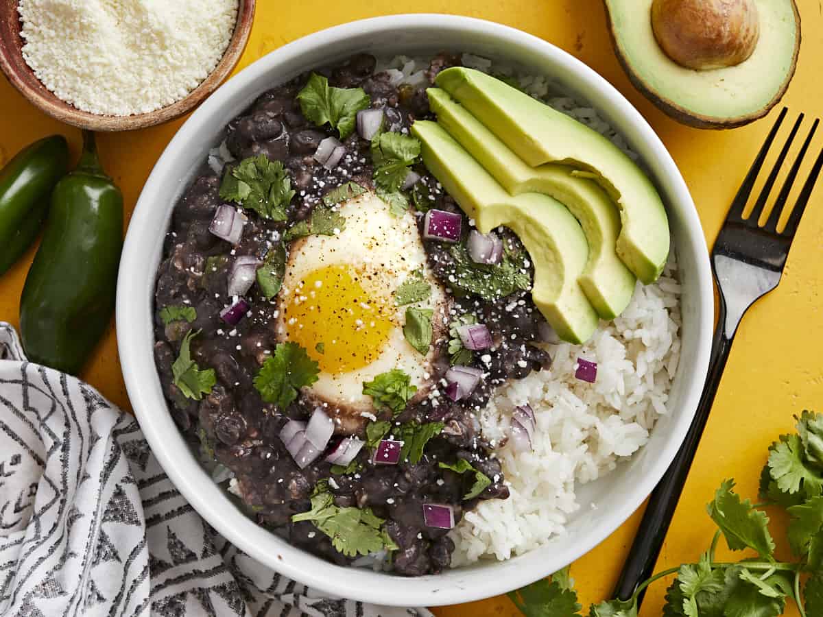 Overhead view of a bowl of black beans with eggs over rice with sliced avocado on the side. 