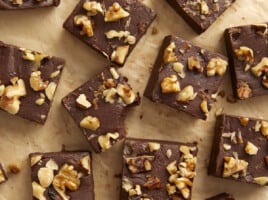 Overhead view of chocolate fudge with walnuts cut into squares on a wooden surface, one with a bite taken out.