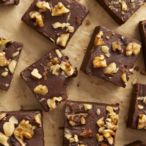 Overhead view of chocolate fudge with walnuts cut into squares on a wooden surface, one with a bite taken out.