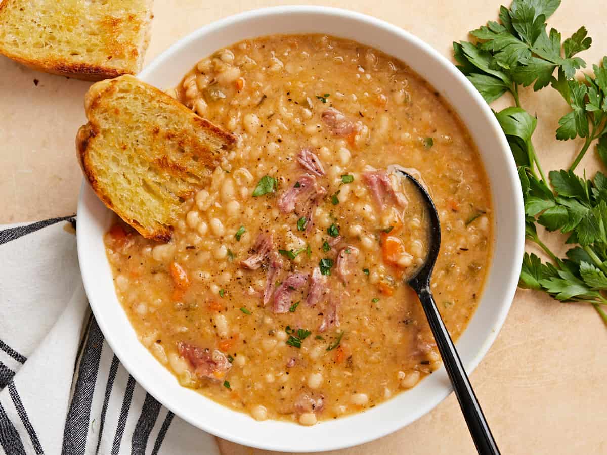 Overhead view of a bowl full of Navy bean soup with crusty bread and a black spoon on the side of the bowl.