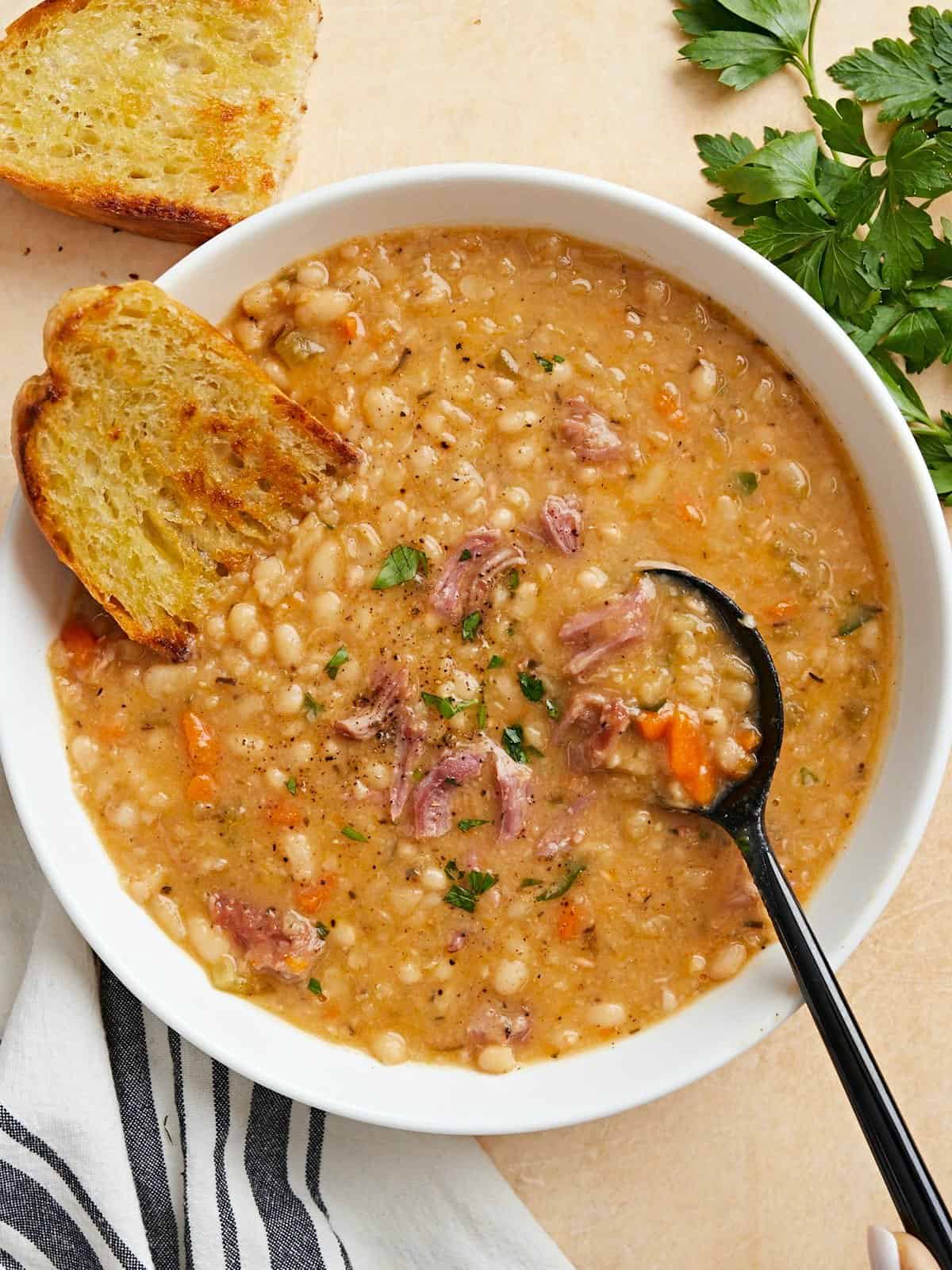 Overhead view of a bowl full of navy bean soup with crusty bread and a black spoon on the side.