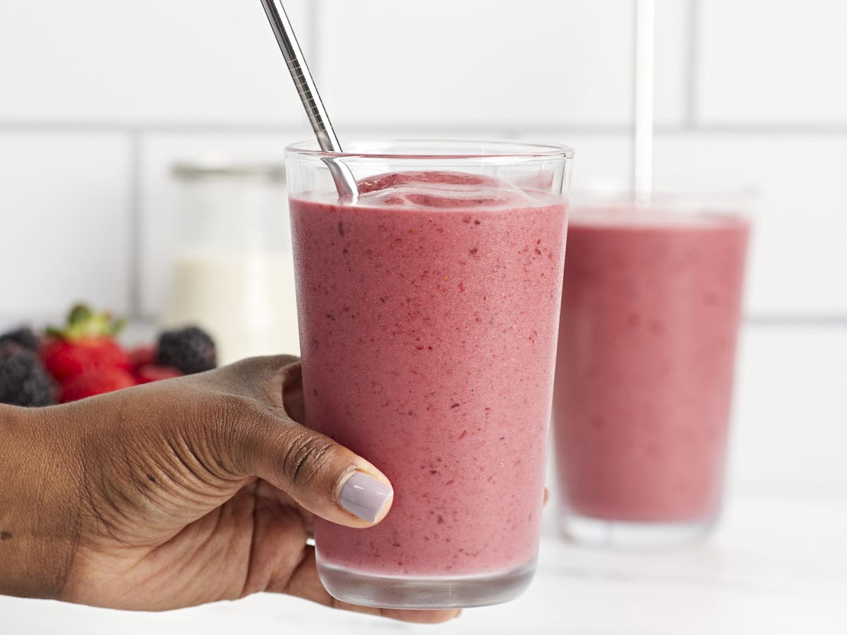 Front side view of two mixed berry smoothies in glasses with one smoothie glass being held by a hand.