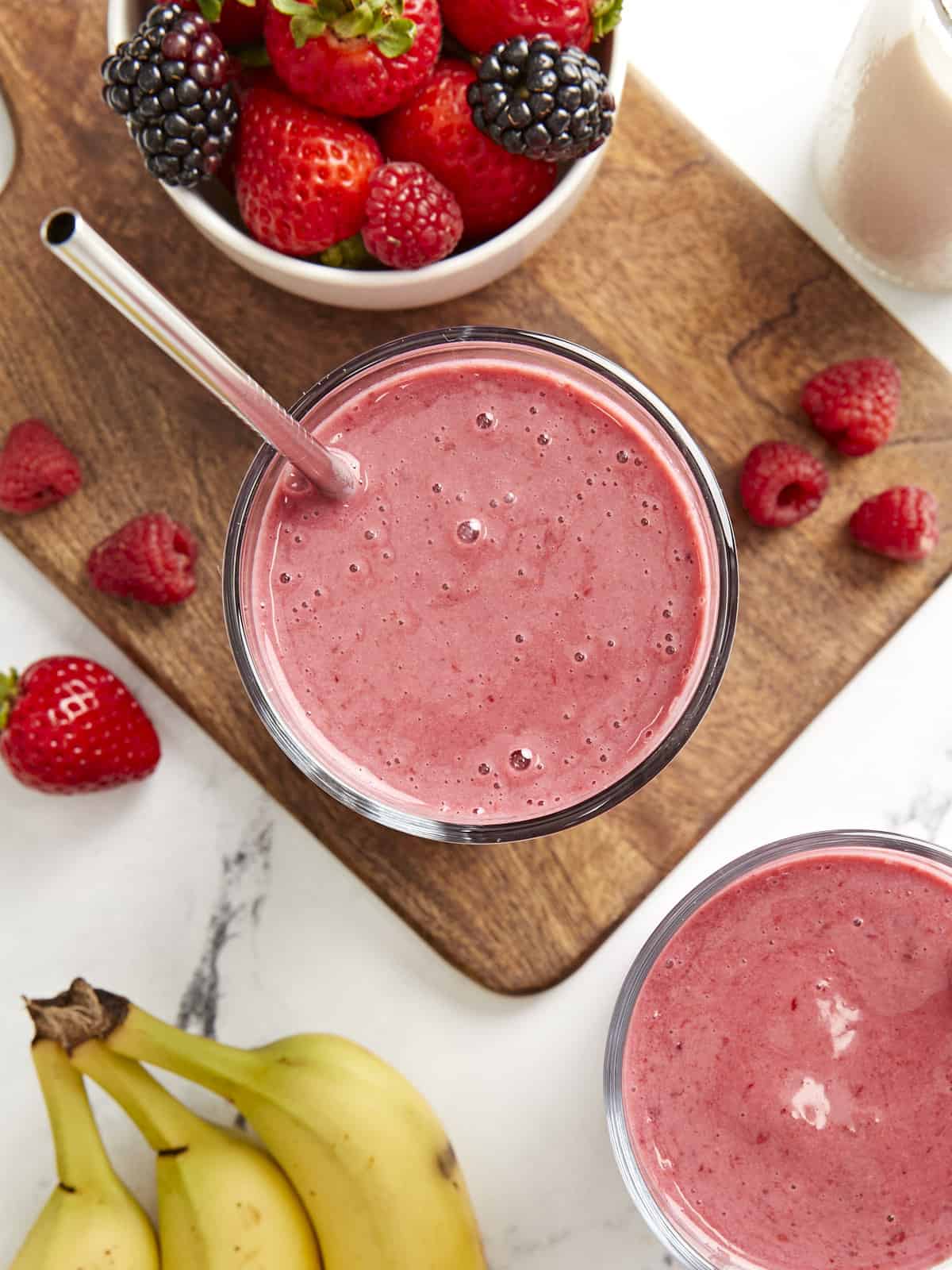 Overhead shot of two mixed berry smoothies in glasses with metal straws and fresh fruit on the side.